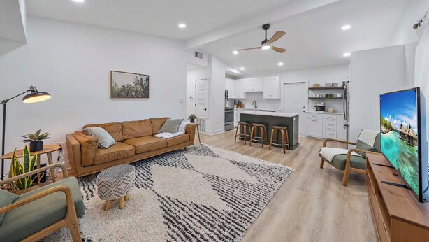 A modern living room with a brown sofa, a coffee table, and a TV. There is a kitchen area with white cabinets and a bar stool. - Memory‑Making Sarasota Stay with Heated Pool, Spa & Modern Comfort (Sarasota)