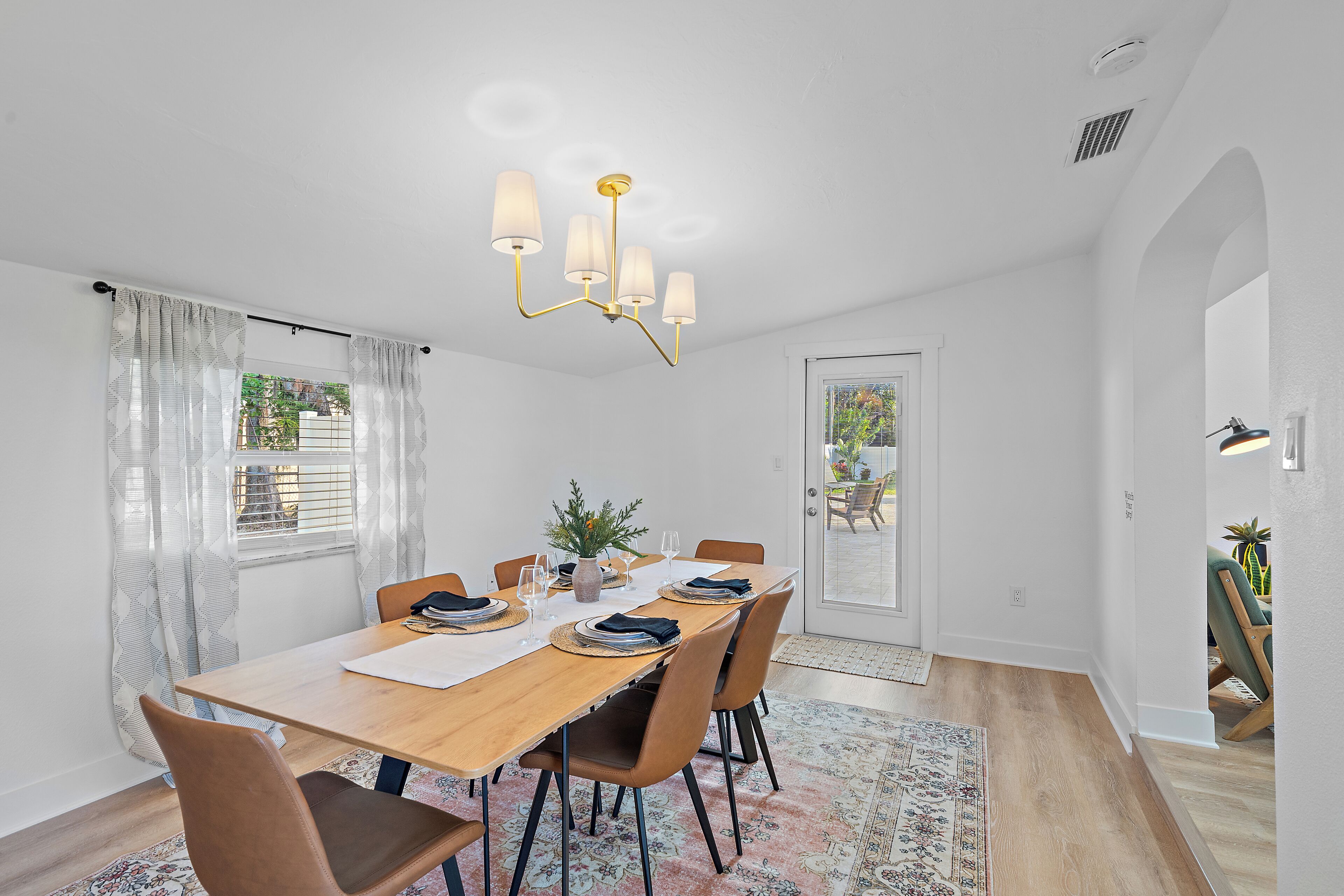 A dining area with a wooden table, brown chairs, a chandelier, and a view through a window with curtains.