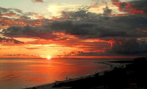 Eine Strandansicht mit Sonnenuntergang über dem Meer, Personen am Ufer und einer fernen Küstenlinie.