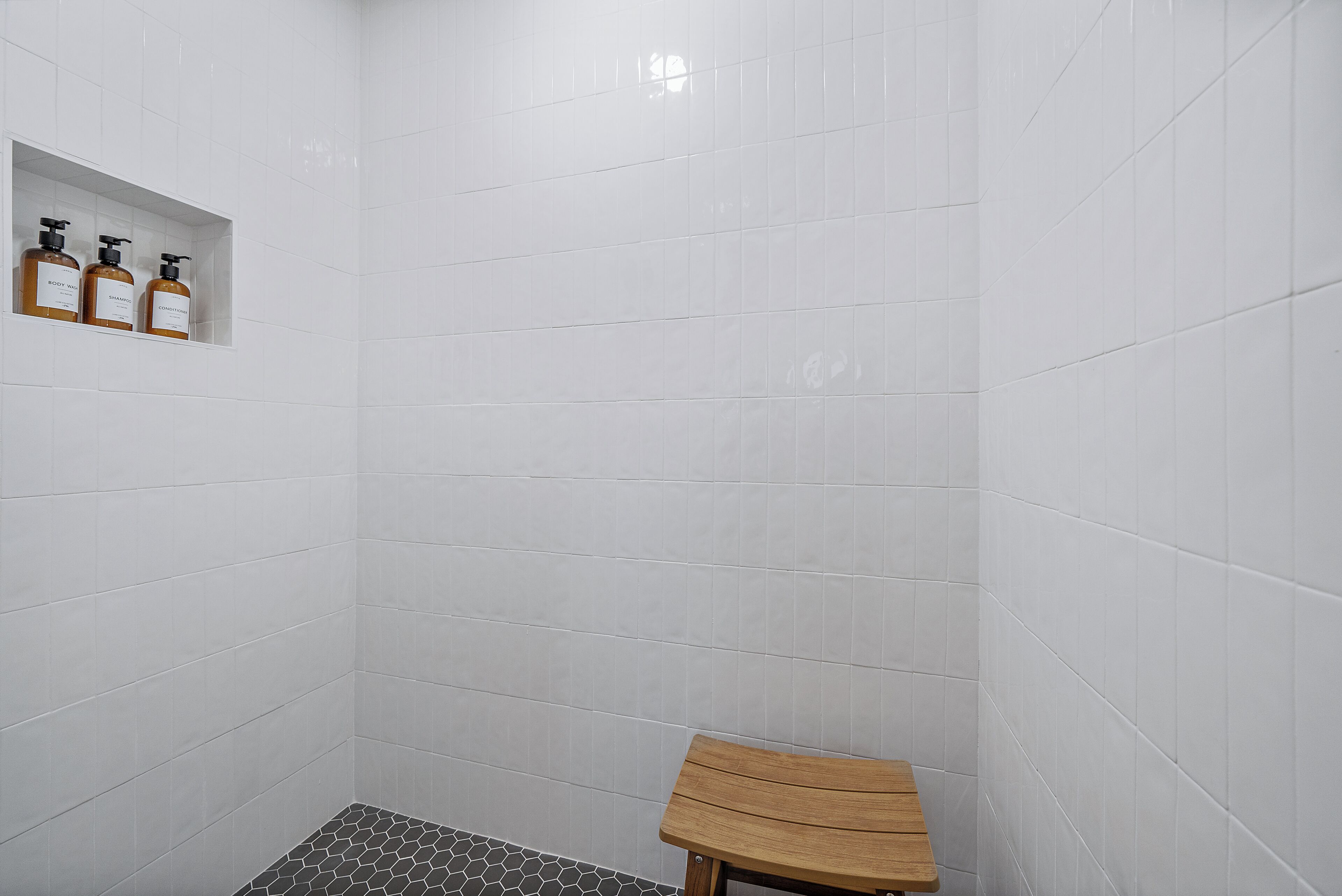 A shower area with white tiles, a wooden bench, and three bottles of shampoo on a shelf.