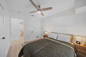 Ein Schlafzimmer mit einem Bett, einem Deckenventilator, einem Nachttisch mit Lampe und einer Tür zu einem anderen Raum.
