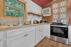 A kitchen with white cabinets, a stainless steel oven, a microwave, and a window with a view of trees. -  Tucker's Hideaway (Morganton)