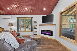 A living room with a fireplace, a sofa, a coffee table, a TV, and a window with a view of a house and trees. -  Tucker's Hideaway (Morganton)
