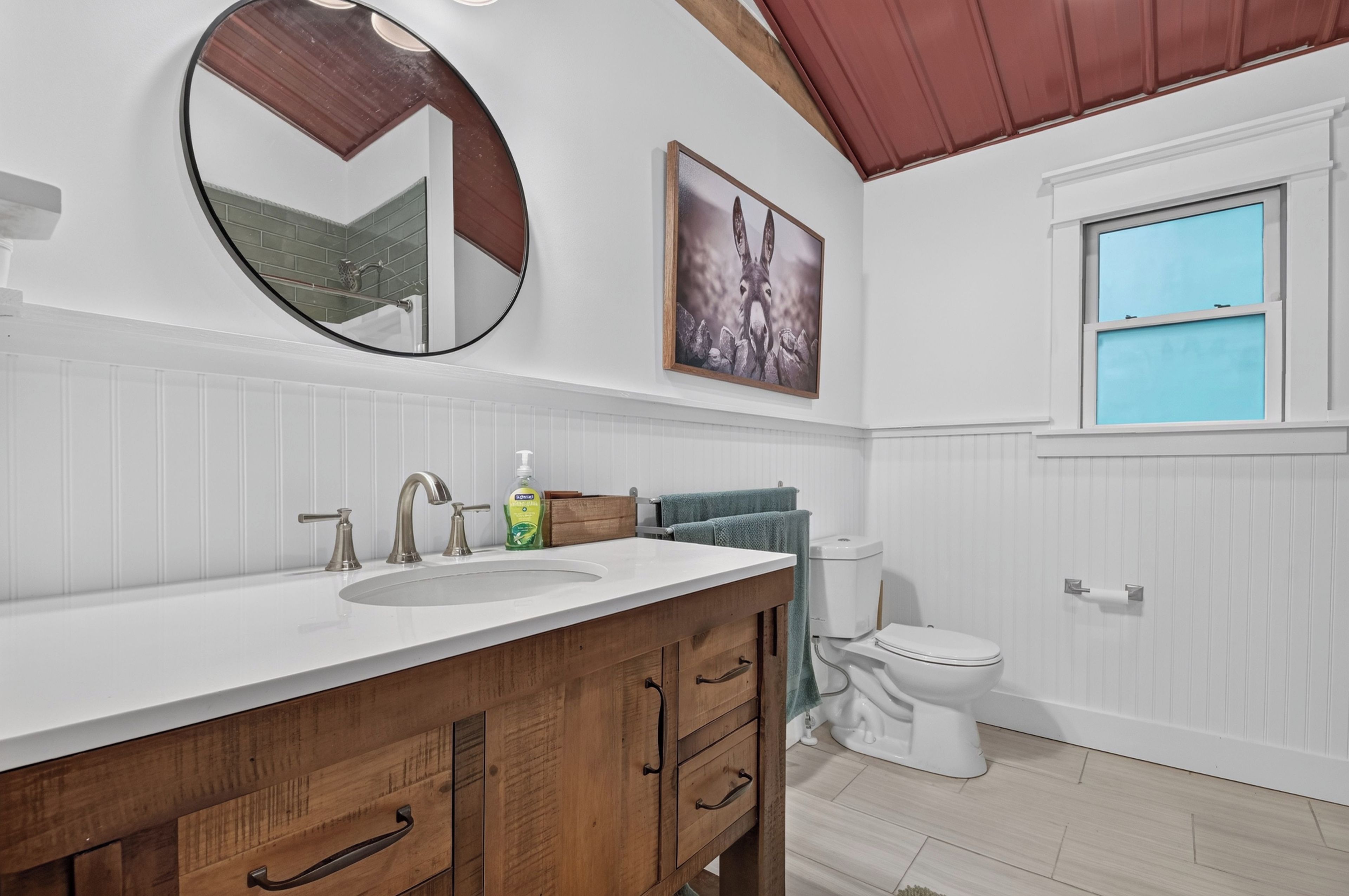 A bathroom with a wooden vanity, a round mirror, a toilet, and a window with a view.