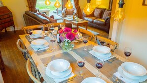 A dining area with a wooden table set for a meal, surrounded by chairs, with a sofa and a view of greenery outside. - Kinloch Lodge - Highland hot tub lodge close to Loch Ness (Drumnadrochit)