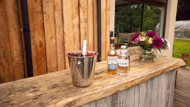 A wooden bar area with a metal bucket containing a bottle, two bottles of alcohol, and a vase with flowers. - Kinloch Lodge - Highland hot tub lodge close to Loch Ness (Drumnadrochit)