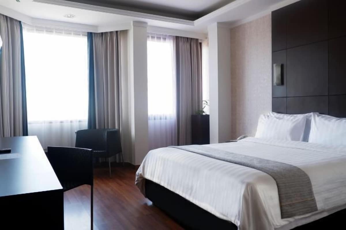 Habitación de hotel con una cama grande, un escritorio y una silla. Hay cortinas en las ventanas y se ve una planta en la esquina.