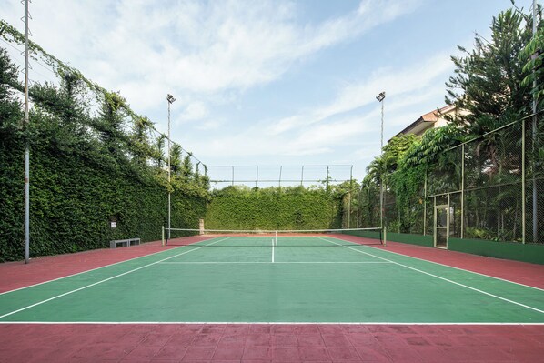 An outdoor tennis court with a green playing surface, surrounded by a chain-link fence, and trees in the background.