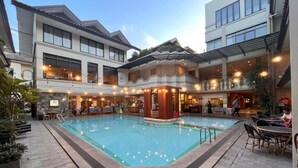 An indoor swimming pool with a wooden deck area, surrounded by buildings with balconies and outdoor seating. - Bentani Hotel & Residences (North Cirebon)