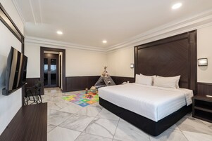 A hotel room with a large bed, a desk, a chair, a television, a wardrobe, and a colorful play mat.