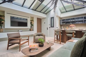 Ein modernes Wohnzimmer mit einem Flachbildfernseher, bequemen Sitzmöglichkeiten, einem Couchtisch mit Büchern und einer Pflanze sowie einer Küchenzeile mit Barhocker und einer Arbeitsplatte.