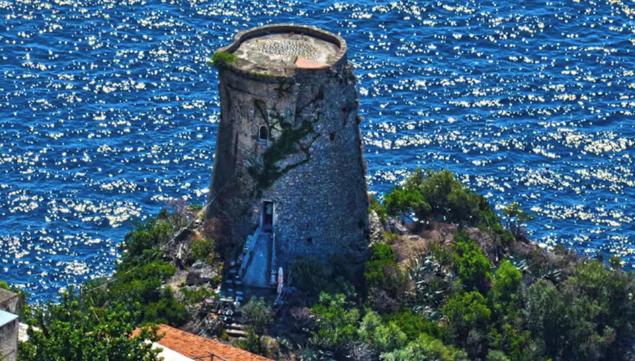 A coastal tower with a staircase leading up to a circular viewing platform, surrounded by greenery and overlooking a sea with boats.