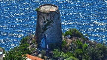 A coastal tower with a staircase leading up to a circular viewing platform, surrounded by greenery and overlooking a sea with boats.