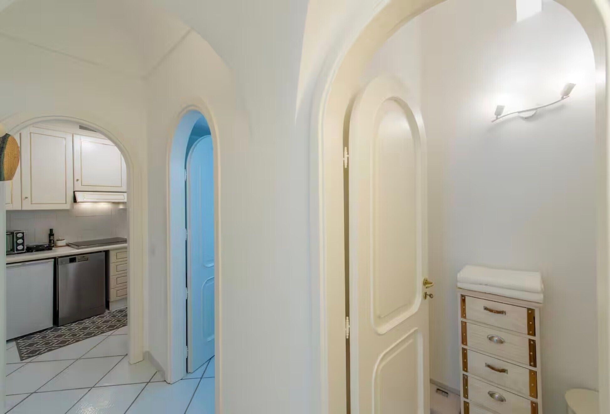 Un corridoio stretto con pareti bianche, una porta blu e un angolo cottura con mobili bianchi e un frigorifero in acciaio inossidabile.