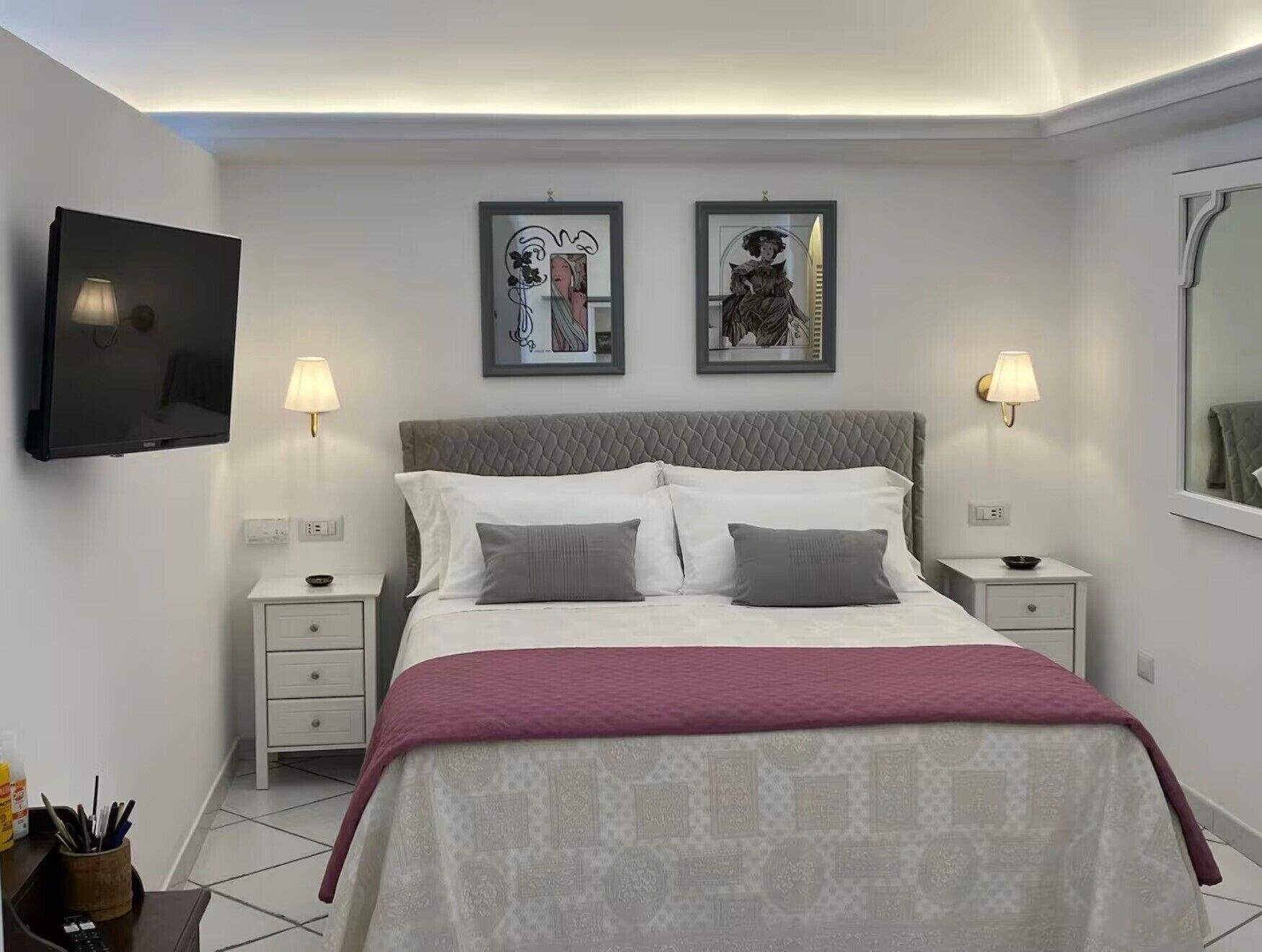 Une chambre d’hôtel comprenant un lit, deux tables de chevet, un téléviseur fixé au mur et deux œuvres d’art encadrées au-dessus du lit.