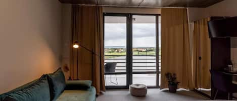 Habitación de hotel con sofá, cama, escritorio, silla, televisor y vista a un cuerpo de agua a través de una puerta corrediza.
