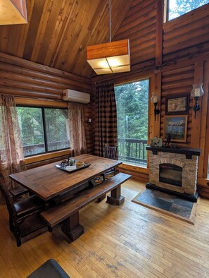 A wooden cabin interior with a dining table, benches, a fireplace, and large windows.