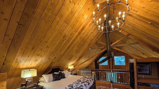 A cozy attic bedroom with a wooden ceiling, a large bed with a patterned bedspread, a nightstand, a lamp, and a view of the outdoors through a window. - Private Mountain cabin, hot tub, 10 min. to Bozeman  10 min. to Bridger Bowl (Bozeman)