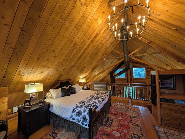A cozy attic bedroom with a wooden ceiling, a large bed with a patterned bedspread, a nightstand, a lamp, and a view of the outdoors through a window.