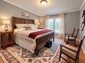 Een slaapkamer met een groot bed, een houten hoofdbord, twee nachtkastjes met lampen, een stoel, een vloerkleed en een raam met gordijnen.