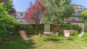 A garden with a table and chairs, surrounded by greenery and a building in the background. - Apartment & Private Garden - Deauville-sevigne (Deauville)