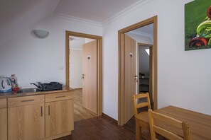 A compact kitchen with a wooden countertop, a kettle, and a sink. There is a wooden dining table with a chair. The room has white walls and a wooden floor.