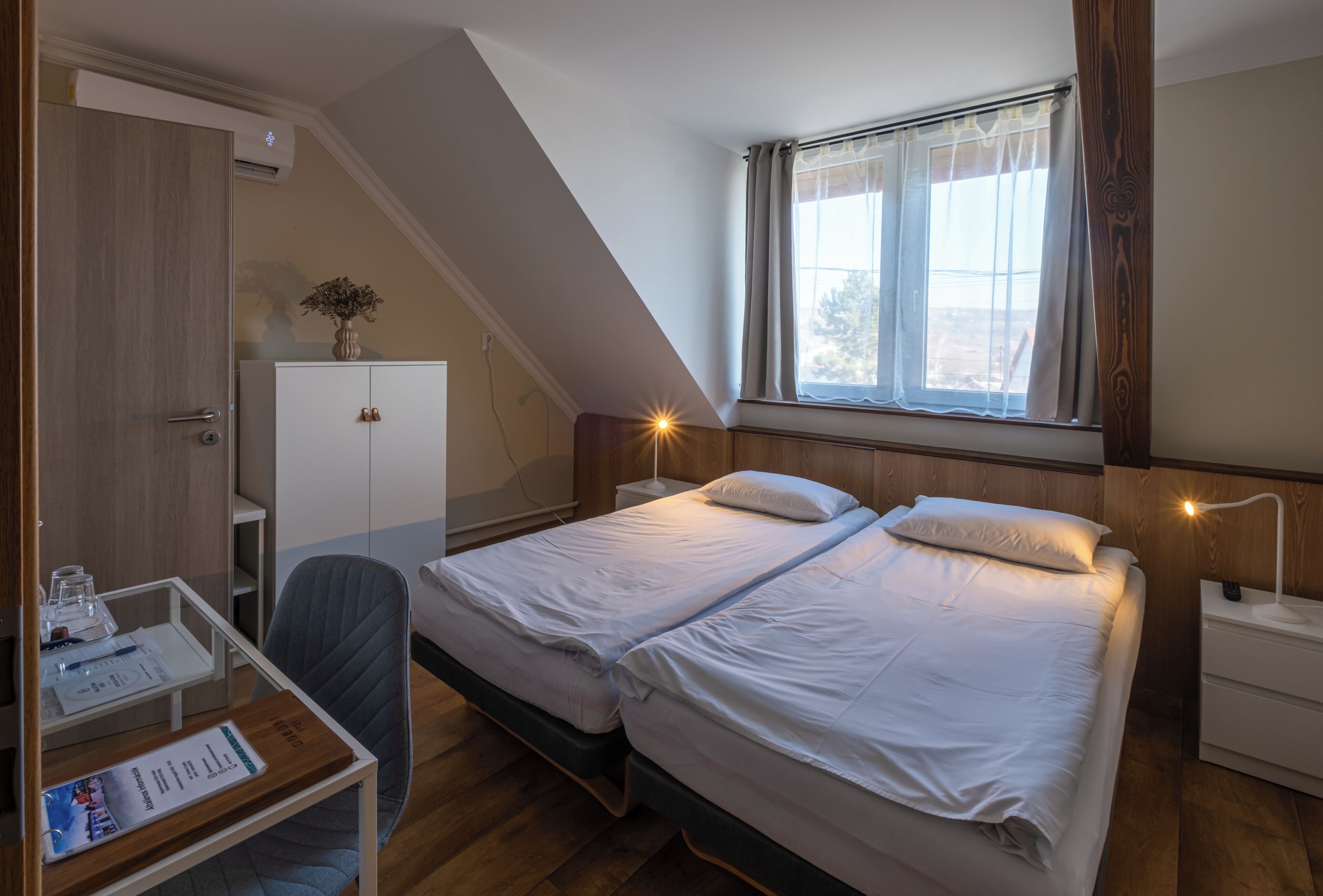 A bedroom with two beds, a desk, a chair, a wardrobe, and a window with curtains.