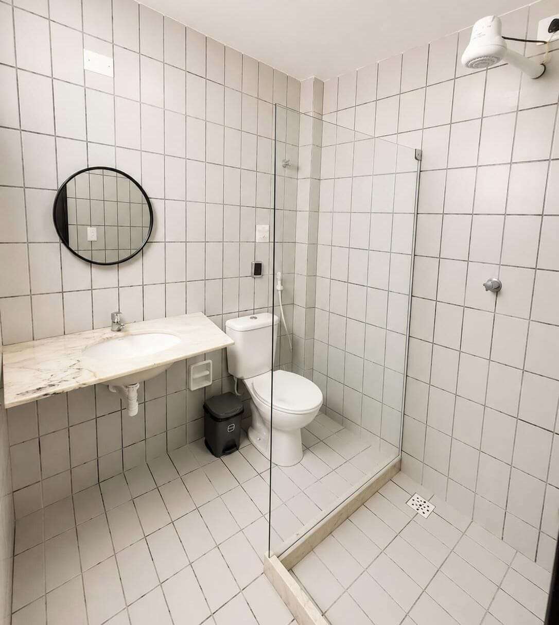 Um banheiro com piso de azulejos brancos, uma pia branca com bancada de mármore, vaso sanitário, box de vidro e um espelho redondo.