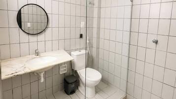 Um banheiro com piso de azulejos brancos, uma pia branca com bancada de mármore, vaso sanitário, box de vidro e um espelho redondo.