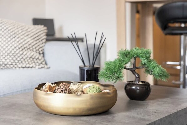 A room with a modern aesthetic, featuring a round, metallic bowl on a table, a bonsai tree in a dark pot, and a black vase with reed sticks.