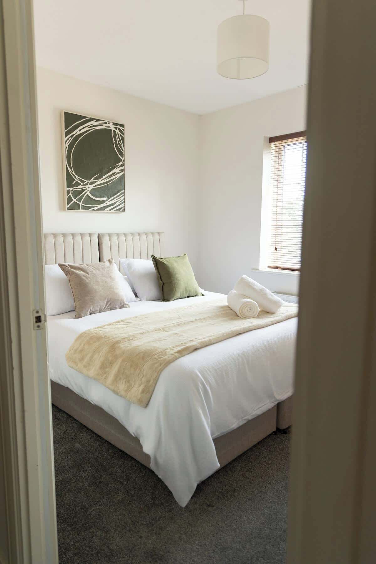 Un dormitorio con cama, almohadas, una colcha, una obra de arte en la pared y una ventana con persianas.