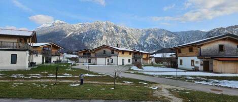 Eine Reihe von Holzhütten mit Bergblick, einem gepflasterten Weg und einer Wiese mit vereinzelten Schneeflecken.