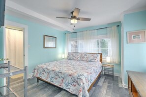 A bedroom with a bed, a ceiling fan, a window with curtains, and a bedside table. - “Captain`s Quarters” at Pirates Perch! (Folly Beach)