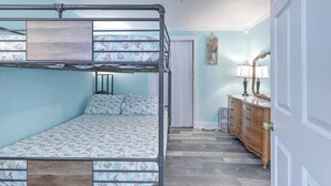 A bunk bed with a patterned bedspread, a dresser with a mirror, and a door in a room with light blue walls. - “Captain`s Quarters” at Pirates Perch! (Folly Beach)