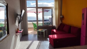 Ein Zimmer mit einem roten Sofa, einem kleinen Tisch, einem an der Wand befestigten Fernseher und einem Fenster mit Meerblick.