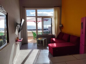A room with a red sofa, a small table, a TV mounted on the wall, and a window with a view of the sea.