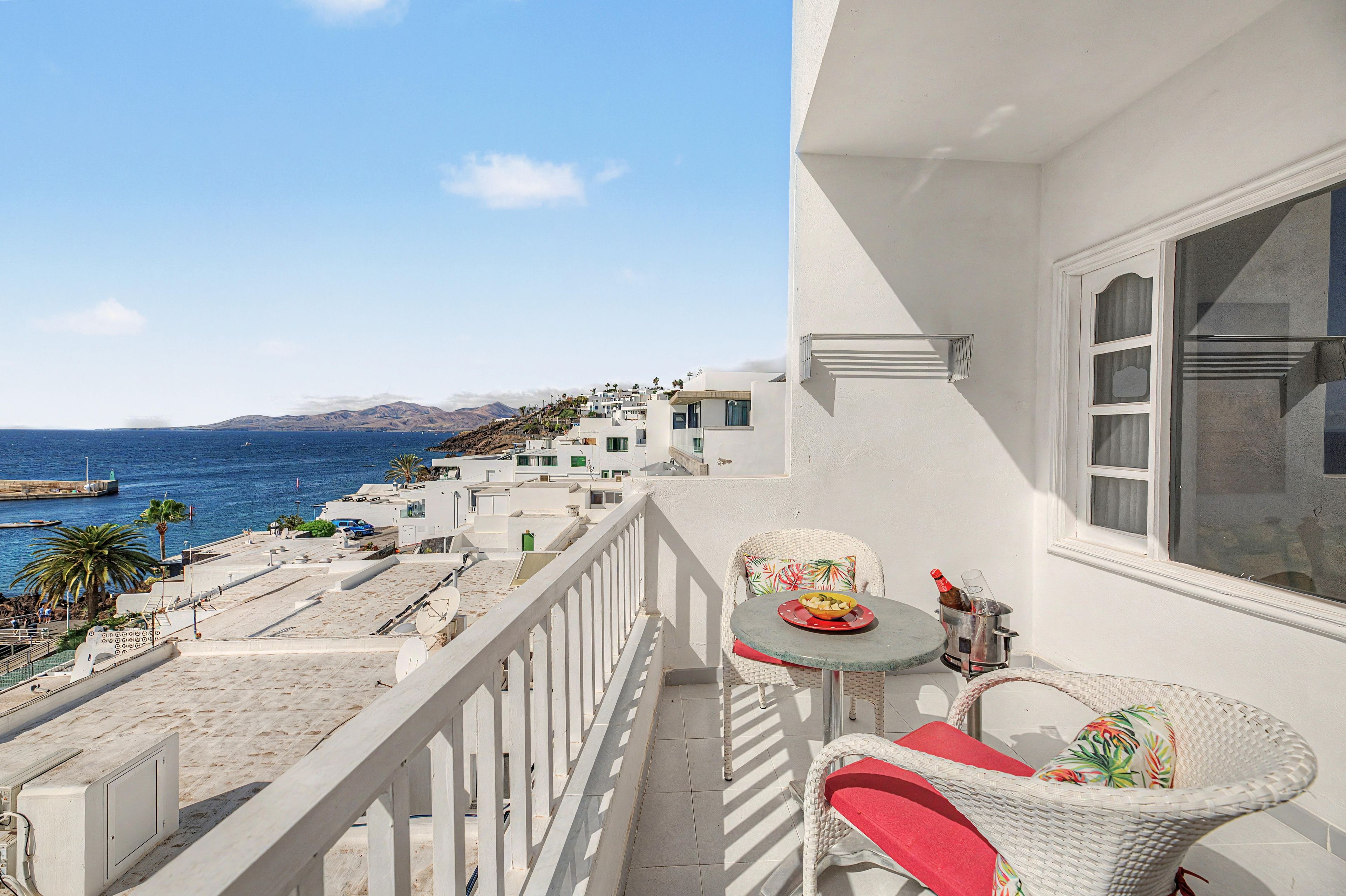 A balcony with a table and chairs, overlooking a coastal area with buildings and the sea.