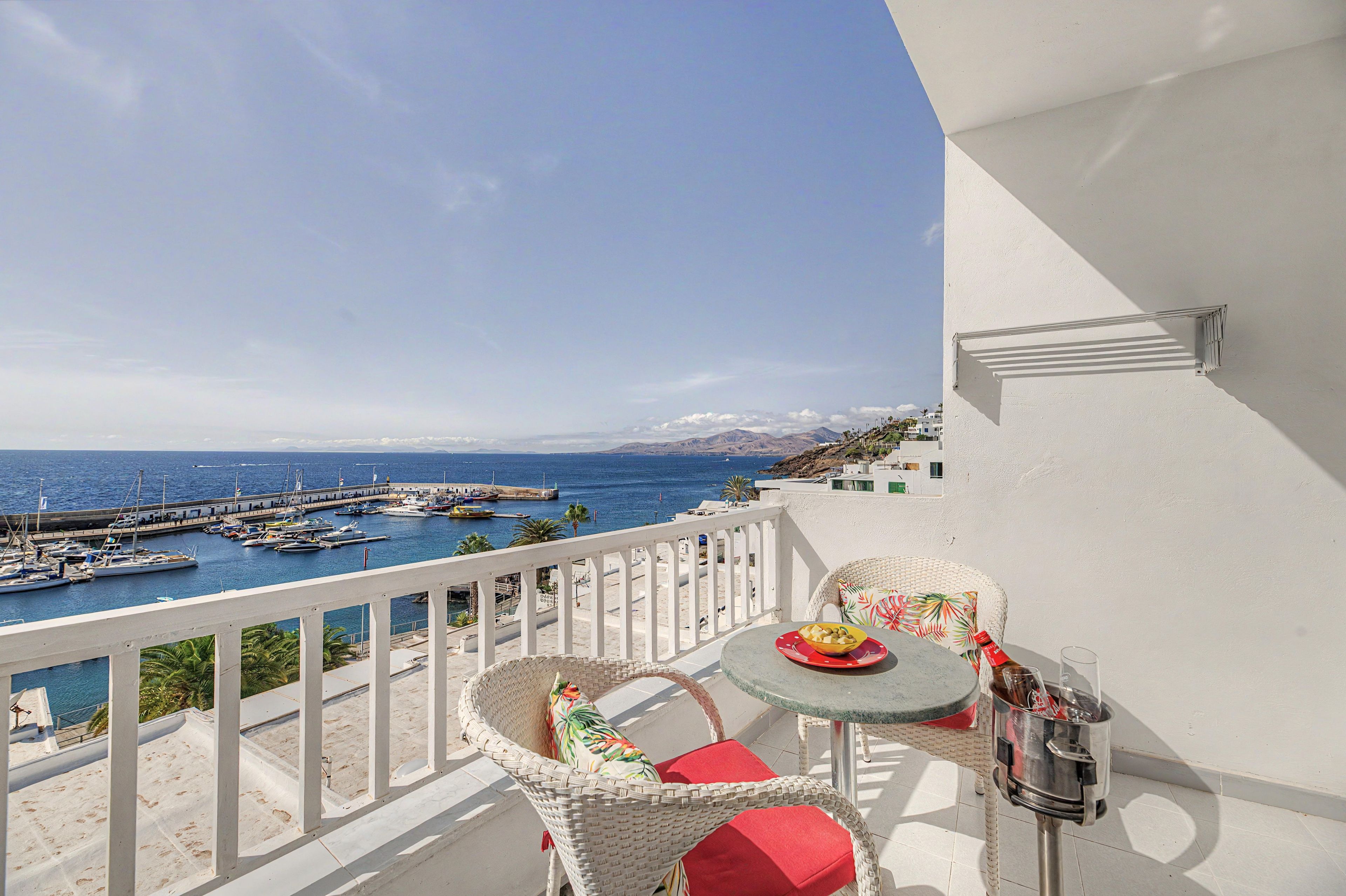 A balcony with a view of a marina and the sea, wicker furniture, and a table with a bottle and glass.