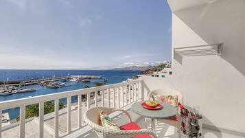 A balcony with a view of a marina and the sea, wicker furniture, and a table with a bottle and glass.