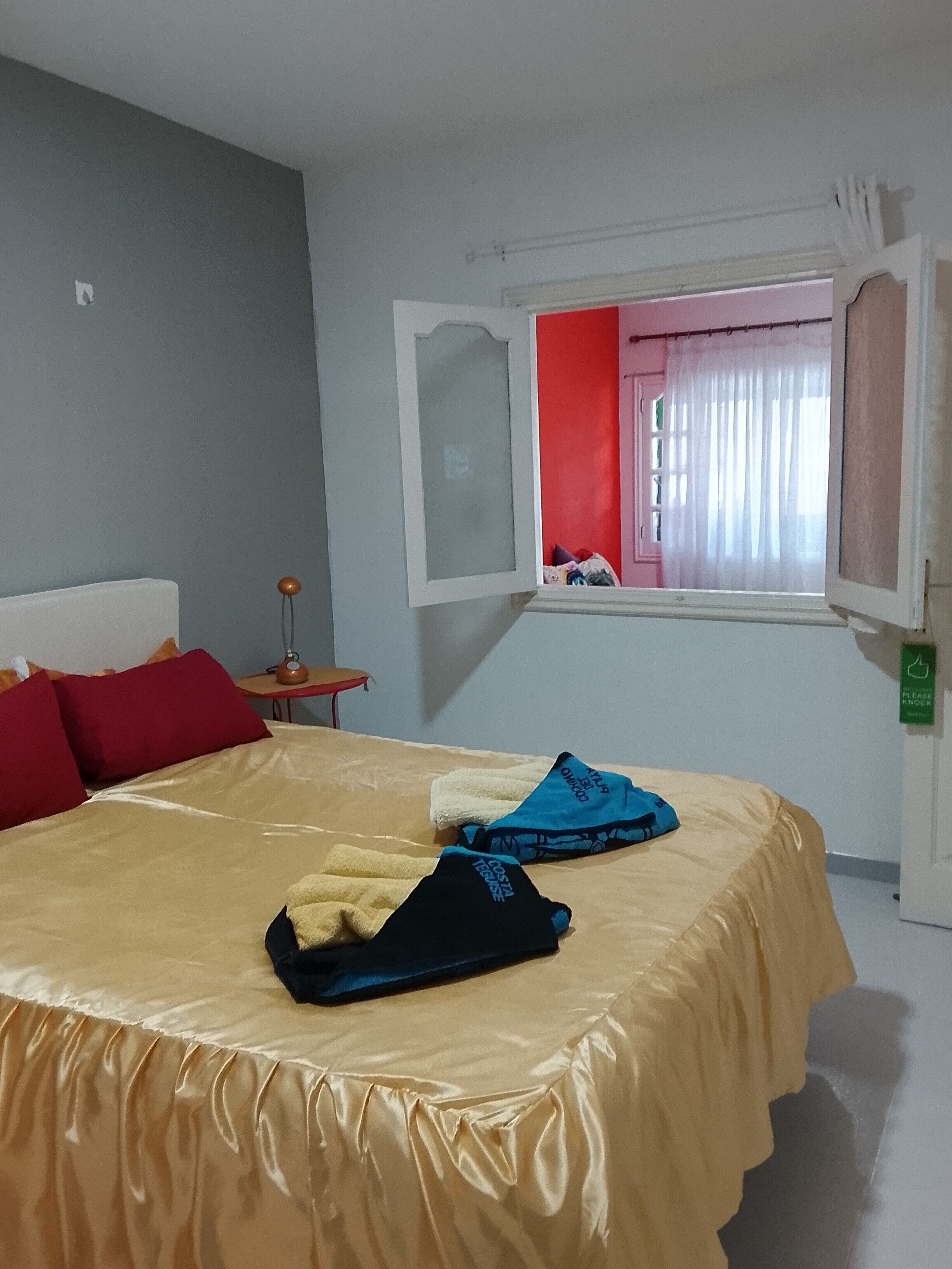 A bed with a beige cover, red pillows, and a window with a view of a red wall and a pink door.