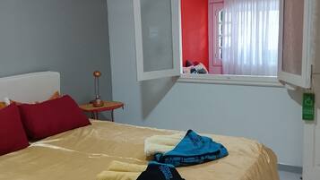 A bed with a beige cover, red pillows, and a window with a view of a red wall and a pink door.
