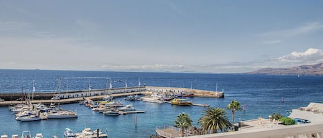 A marina with boats docked, a pier extending into the water, and a clear view of the sea and distant mountains.