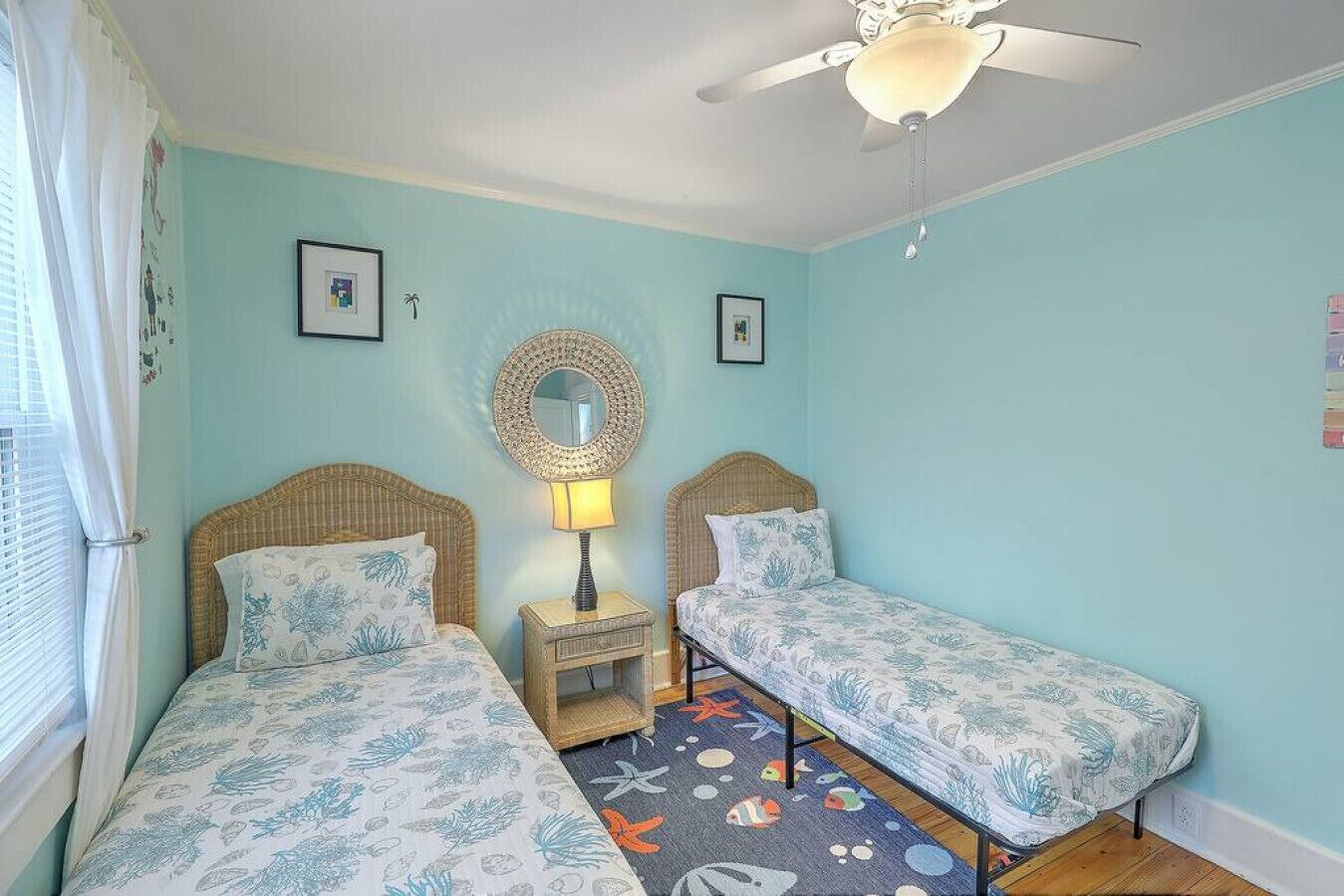 A room with two beds, a ceiling fan, a round mirror, and a small table with a lamp.