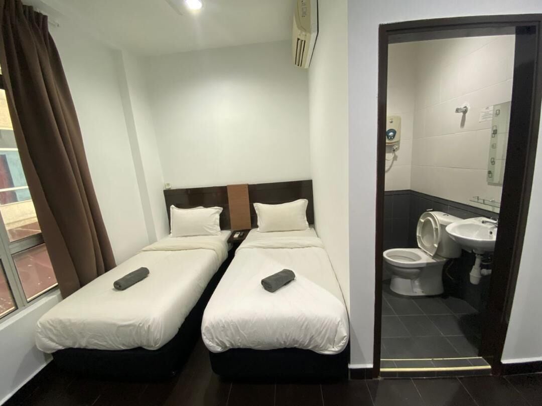 Une chambre d’hôtel avec deux lits simples, une fenêtre avec des rideaux et une salle de bain équipée de toilettes et d’un lavabo.