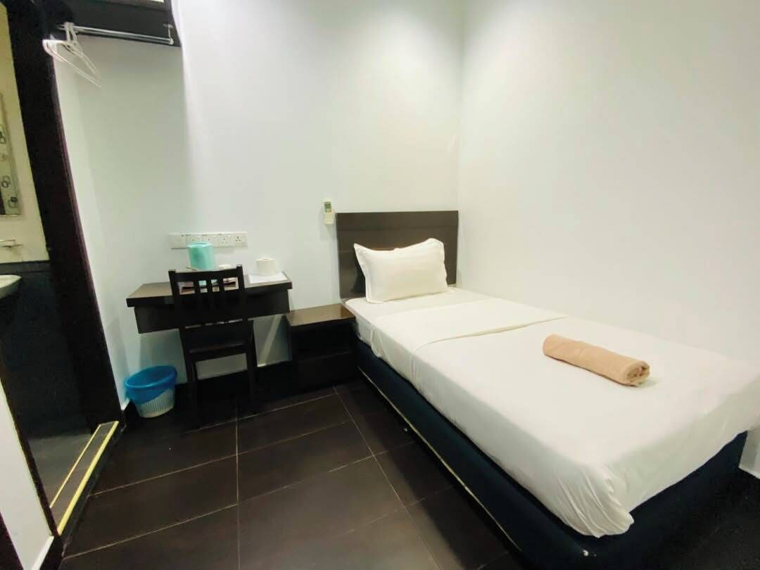 Une petite chambre avec un lit simple, un bureau, une chaise et une poubelle bleue.