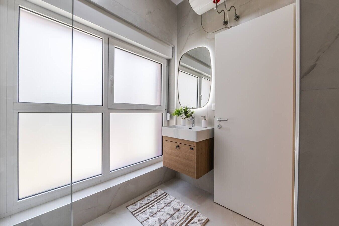 A modern bathroom with a large window, a mirror, a sink with a wooden vanity, and a door.