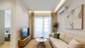 Una sala moderna con sofá, televisor sobre un mueble de madera, una mesa redonda y un ventanal grande con cortinas.