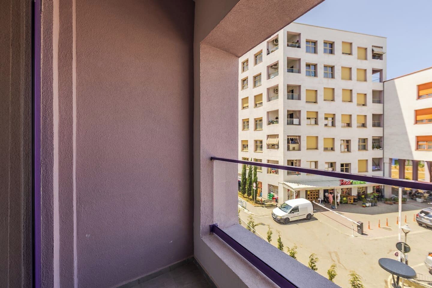 Un balcón con vista a un edificio de varios pisos, una calle con coches estacionados y una pequeña zona de estar al aire libre.
