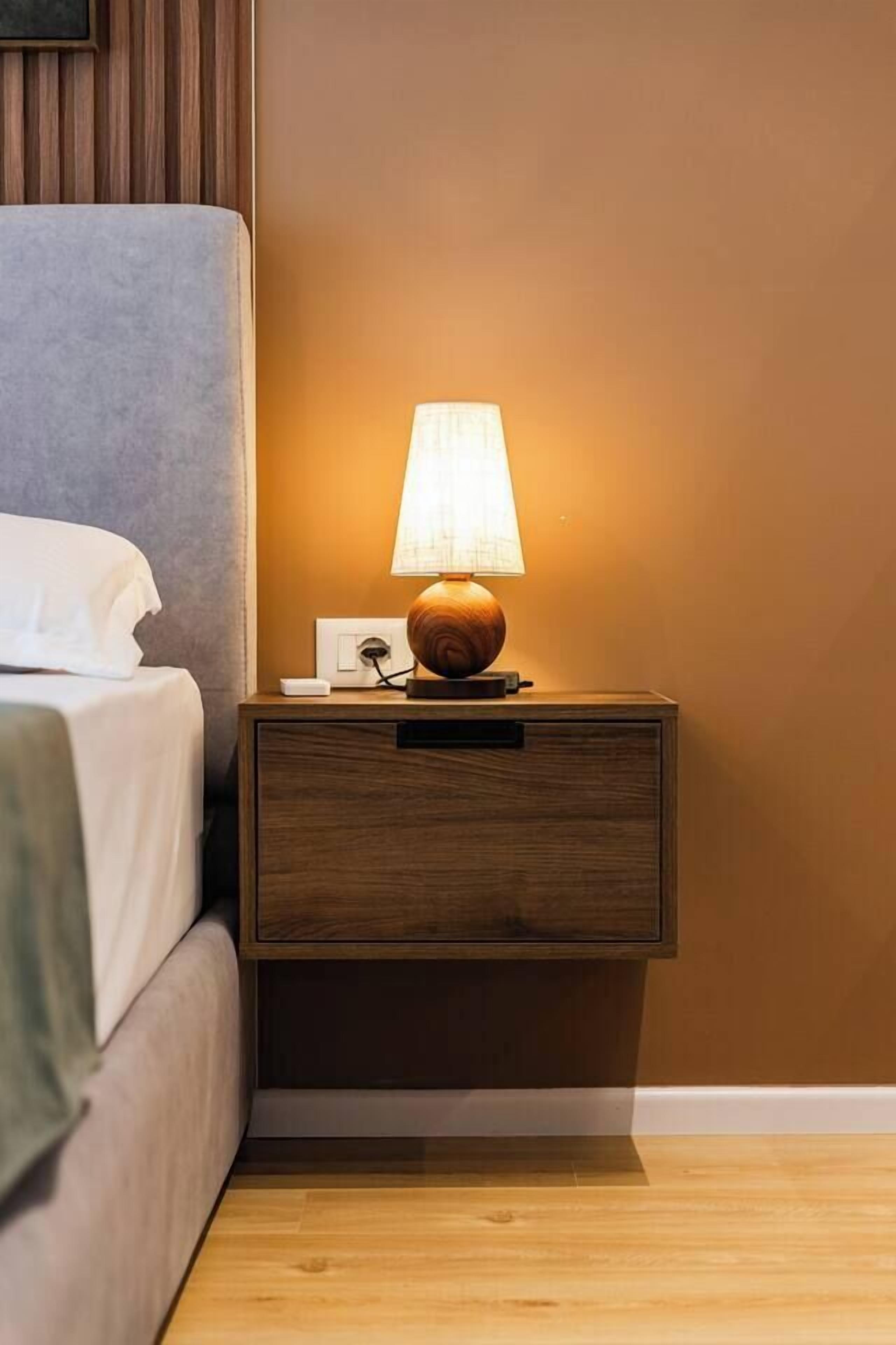 A wooden bedside table with a lamp, a bed with white bedding, and a wall with a wooden panel design.