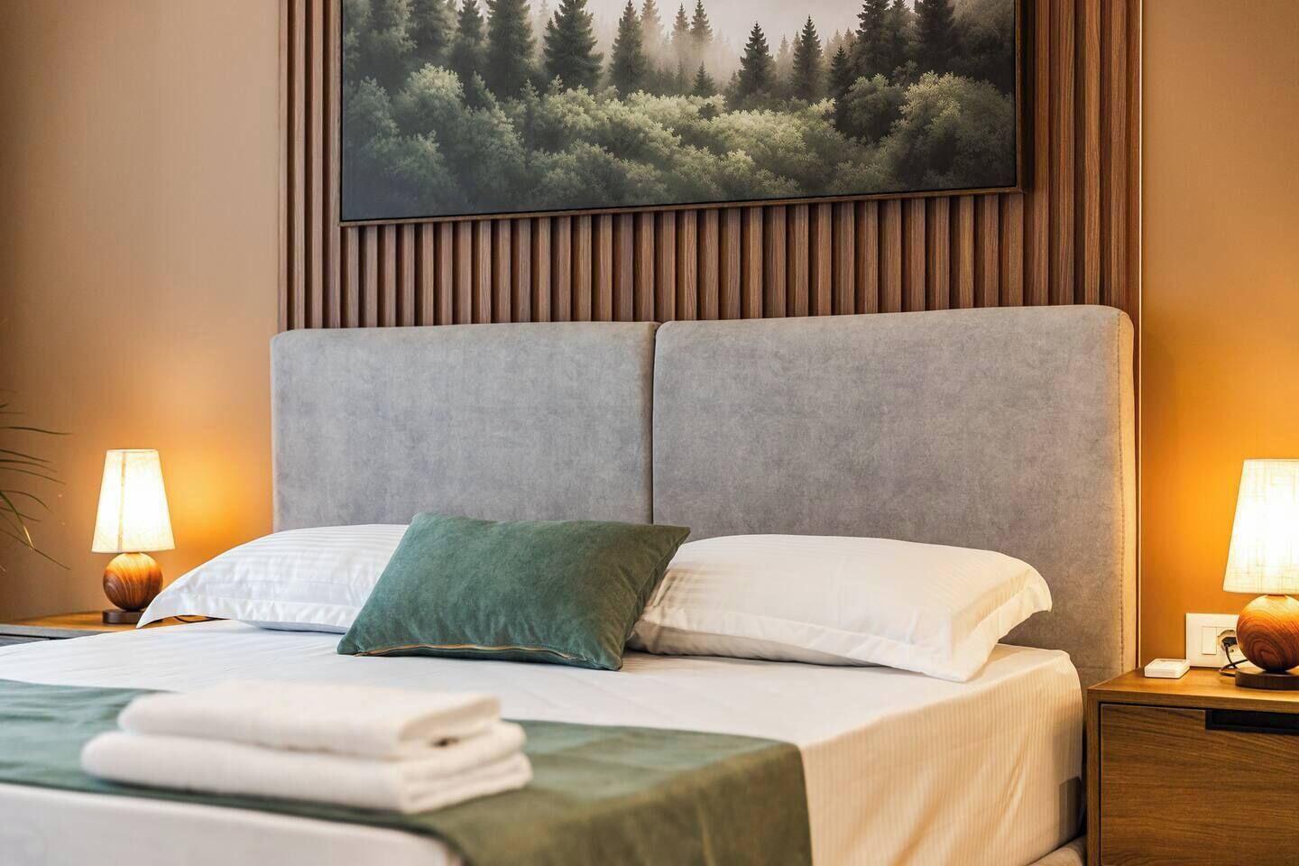 A neatly made bed with a grey headboard, two pillows, and a green throw pillow. There are two bedside lamps on wooden nightstands, and a painting of a forest on the wall.